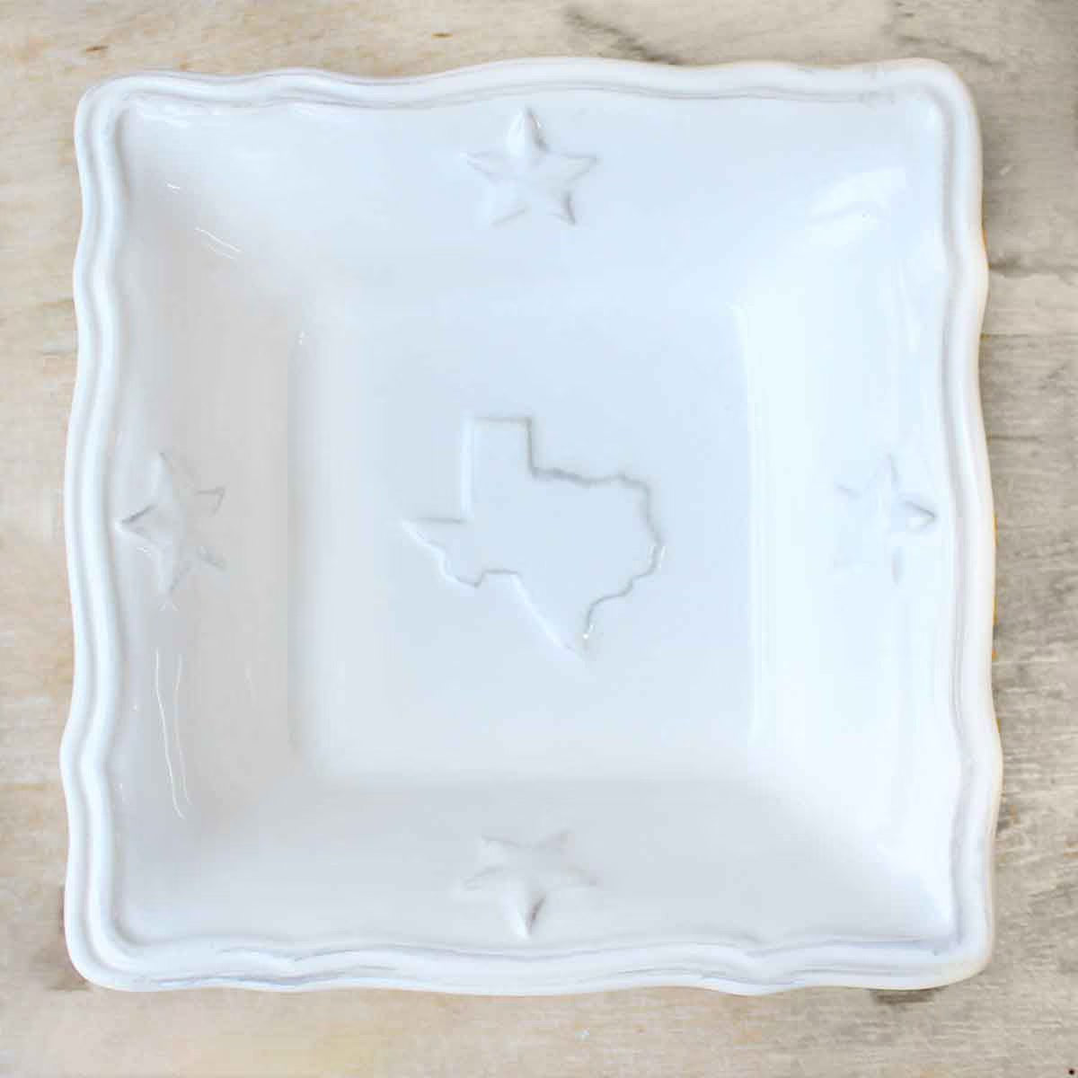 The Texas Tidbit Dish is a square white ceramic plate with embossed stars at each corner and a central Texas outline. Its wavy edge highlights its dolomite composition. Dishwasher and microwave safe. Available sizes not specified.