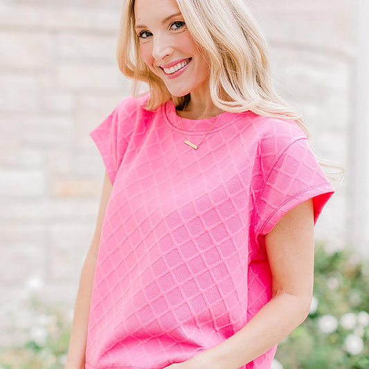 A smiling woman with blonde hair, wearing a bright pink "Ella" t-shirt and a delicate necklace, standing against a soft-focus beige brick wall.