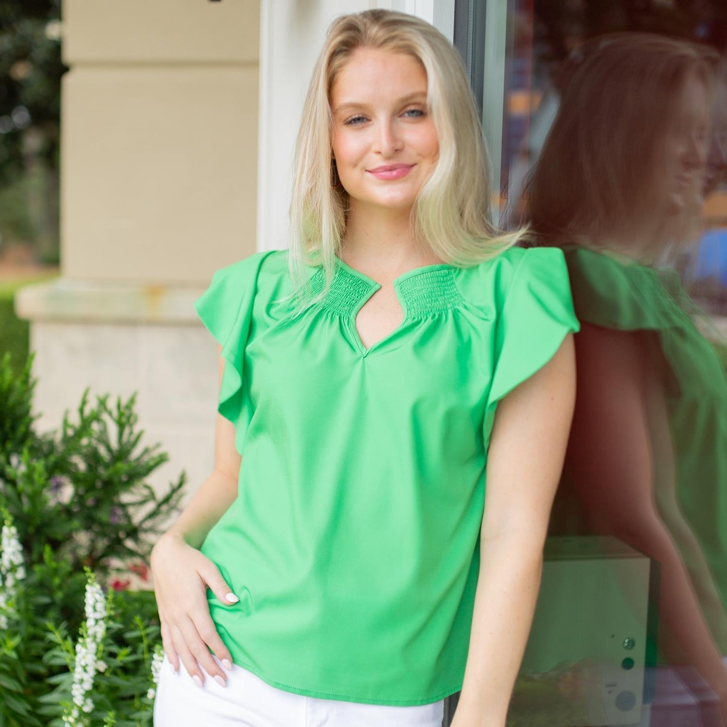 A confident woman in a Penelope Top and white pants, with appropriate sizing, leans against a glass window with her reflection visible, hands in pockets, smiling gently.