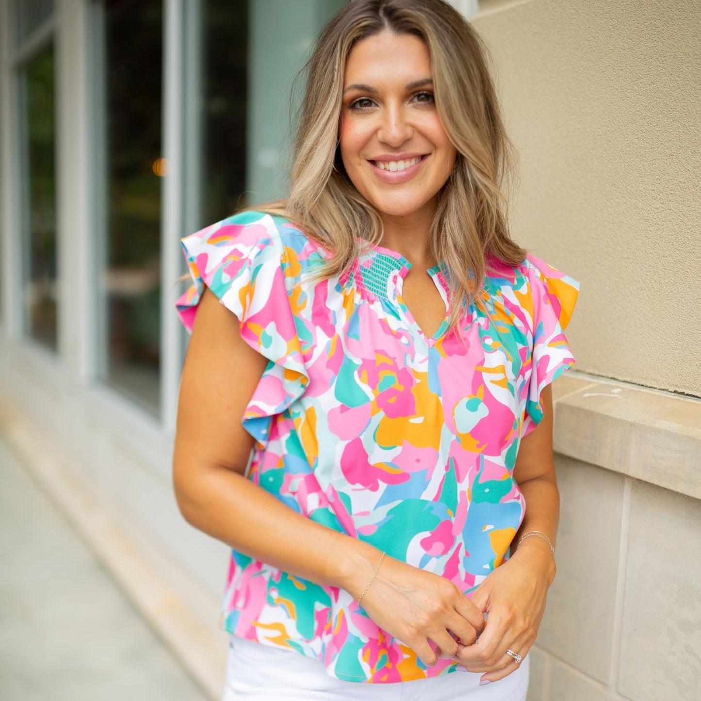 A smiling woman with shoulder-length hair, wearing a Penelope Top with puffed sleeves and white pants, stands outside.