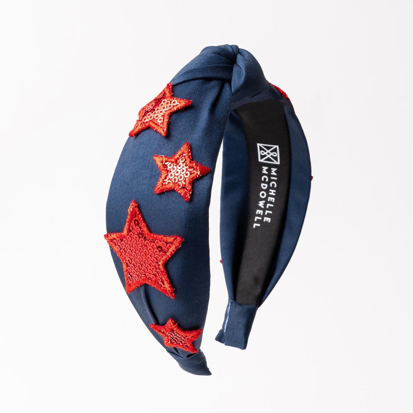 The Emma Headband features a navy blue fabric adorned with red sequined stars and a stylish twisted knot design on a white backdrop. Available in one size.