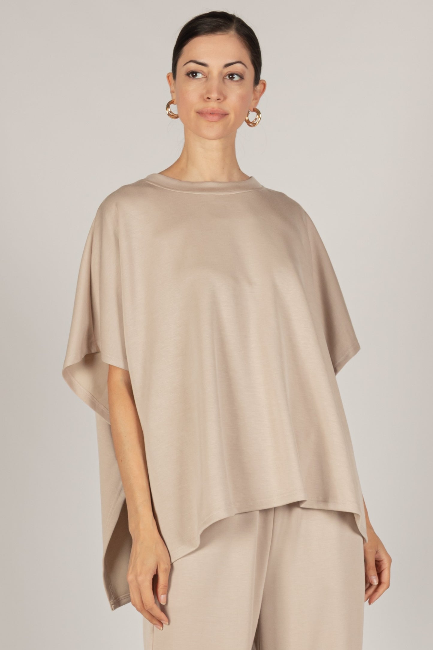 A woman in a P. CILL Butter Modal Side Slit Cape Top with sizing details and matching pants, stands with her hair pulled back and hoop earrings.