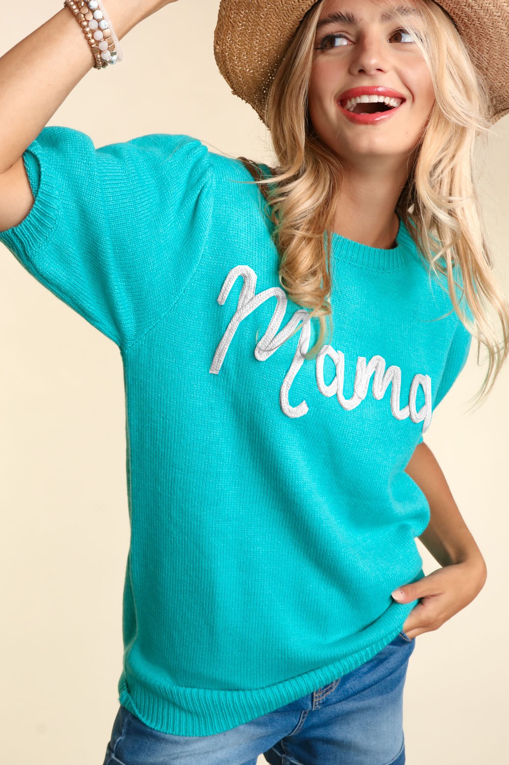 A cheerful woman wearing a vibrant teal Mama Embroidered Pop Letter Short Bubble Sleeve Sweater - Plus, a straw hat, and denim jeans, joyfully posing in a studio setting.