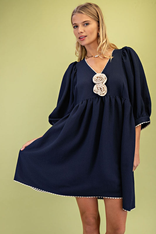 Navy blue V-neck mini dress with puffed sleeves and ric-rac trim. Features two cream-colored rosettes on the chest. Available in sizes XS to XL.