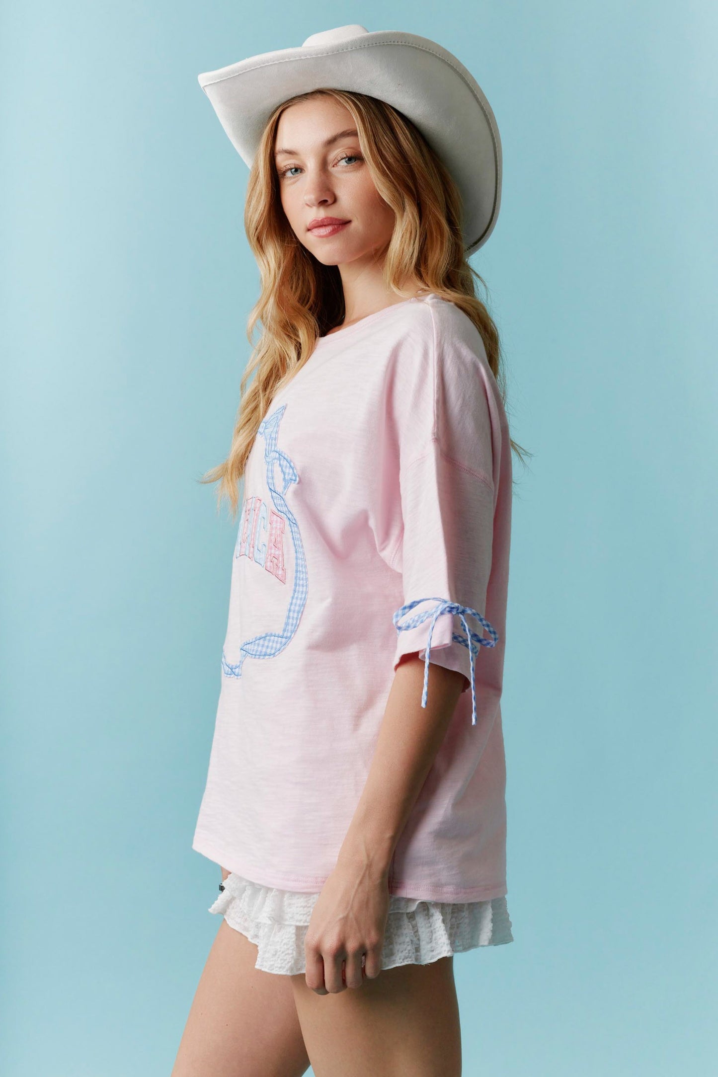 Americana Gingham Bow T-Shirt: White short-sleeve tee with a red and blue gingham bow graphic on the chest. Sizes available: XS, S, M, L, XL. White Ruffled Shorts: High-waisted white shorts featuring layered ruffle detailing at the hem. Sizes available: XS, S, M, L, XL. White Cowboy Hat: Classic western-style hat in white with a structured brim. One size fits most.