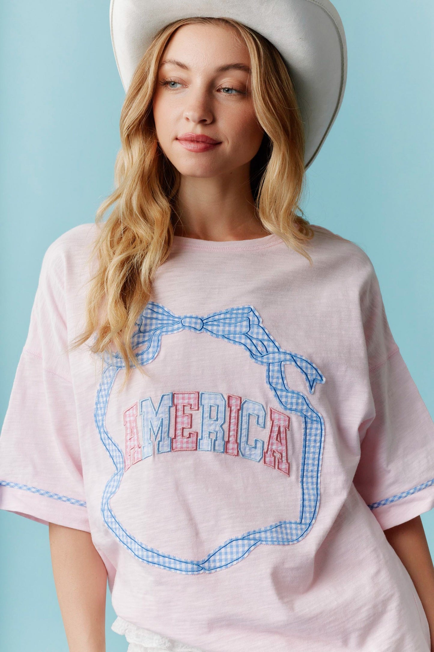 Americana Gingham Bow T-Shirt: White tee with “AMERICA” in red, white, and blue lettering and a gingham bow outline. Sizes available: S, M, L, XL.