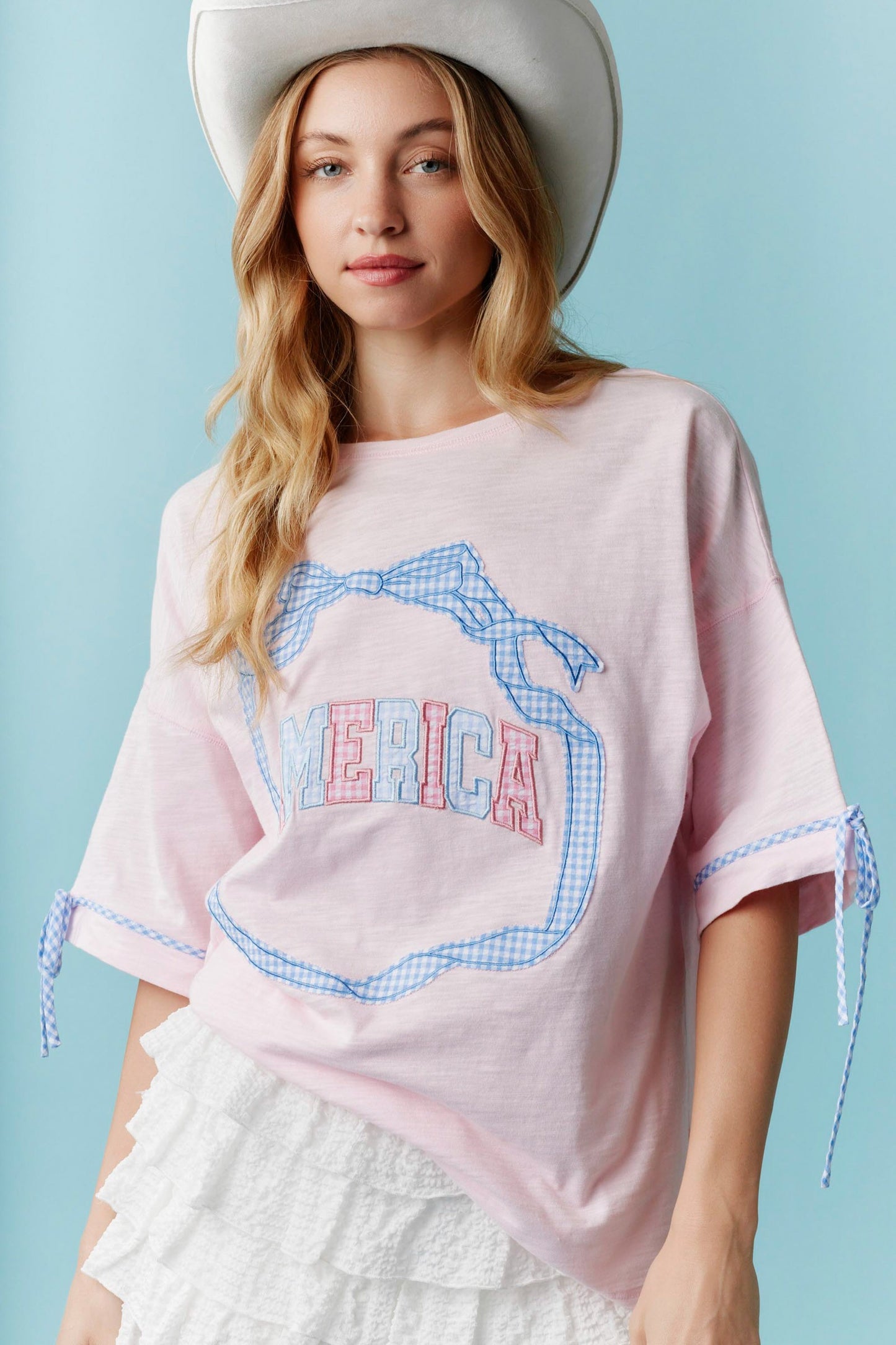 Americana Gingham Bow T-Shirt features blue gingham trim, bold “MERICA” lettering, and a bow accent. Sizes available: S, M, L, XL.