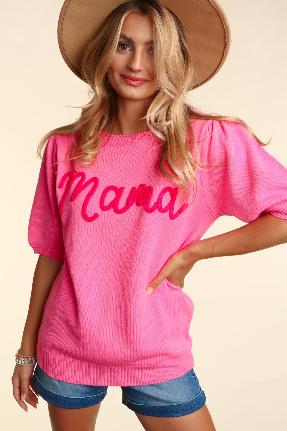 A woman wearing a pink Mama Embroidered Pop Letter Short Bubble Sleeve Sweater, denim shorts, and a wide-brimmed hat, smiling subtly at the camera.