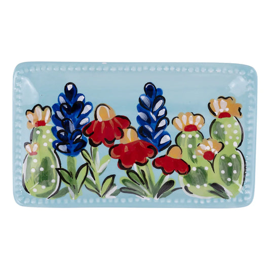 The Texas Wildflower Trinket Tray is a rectangular, hand-painted ceramic plate with a light blue background adorned with colorful flowers and cacti in red, blue, green, and yellow. The plate's border features small white dots. Available sizes: Small, Medium.