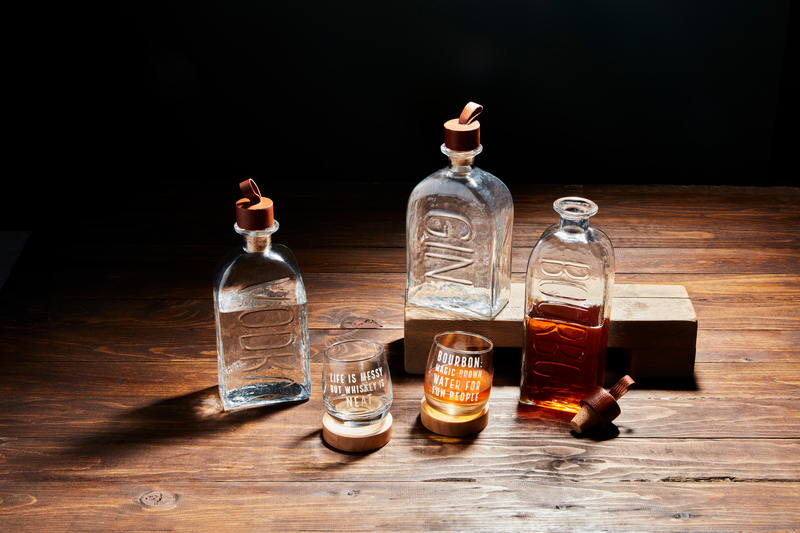 Four embossed glass decanters, one half-filled with amber liquid and three empty with cork stoppers. Two empty glasses, one marked "LOVE IS MESSY // BUY WHISKEY," sit in front. Sizes: Standard decanter size.