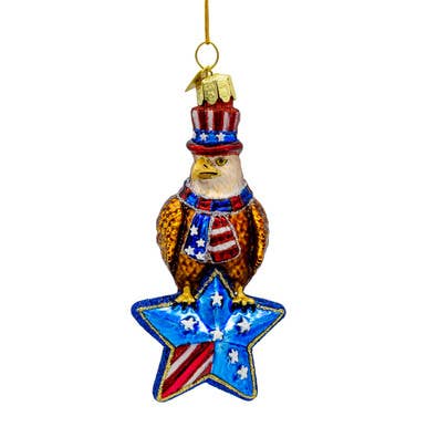 The Noble Gems Glass American Eagle Ornament is a glass eagle with a red, white, and blue hat and scarf, perched on a blue star with stripes and white stars. Hangs by a gold string. Sizes available: 5 inches.