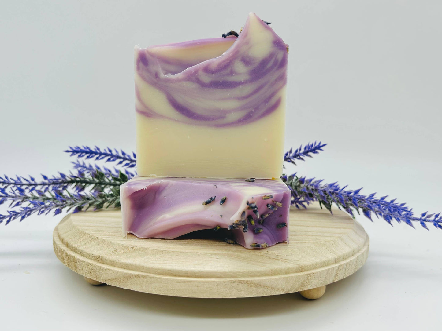 This product includes two Lavender Handmade Soap Bars that feature artistic swirls of purple and white. The soap bars are presented on an elegant wooden dish and further adorned with natural lavender sprigs.