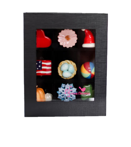 The Keepsake Box Holds 9 features a black exterior, a transparent lid, and a magnetic closure. It beautifully displays nine colorful chocolates arranged in a 3x3 grid. Includes shapes like a red heart, pink flower, Santa hat, American flag, nest with eggs, and beach ball. Available Size: Holds 9 chocolates.