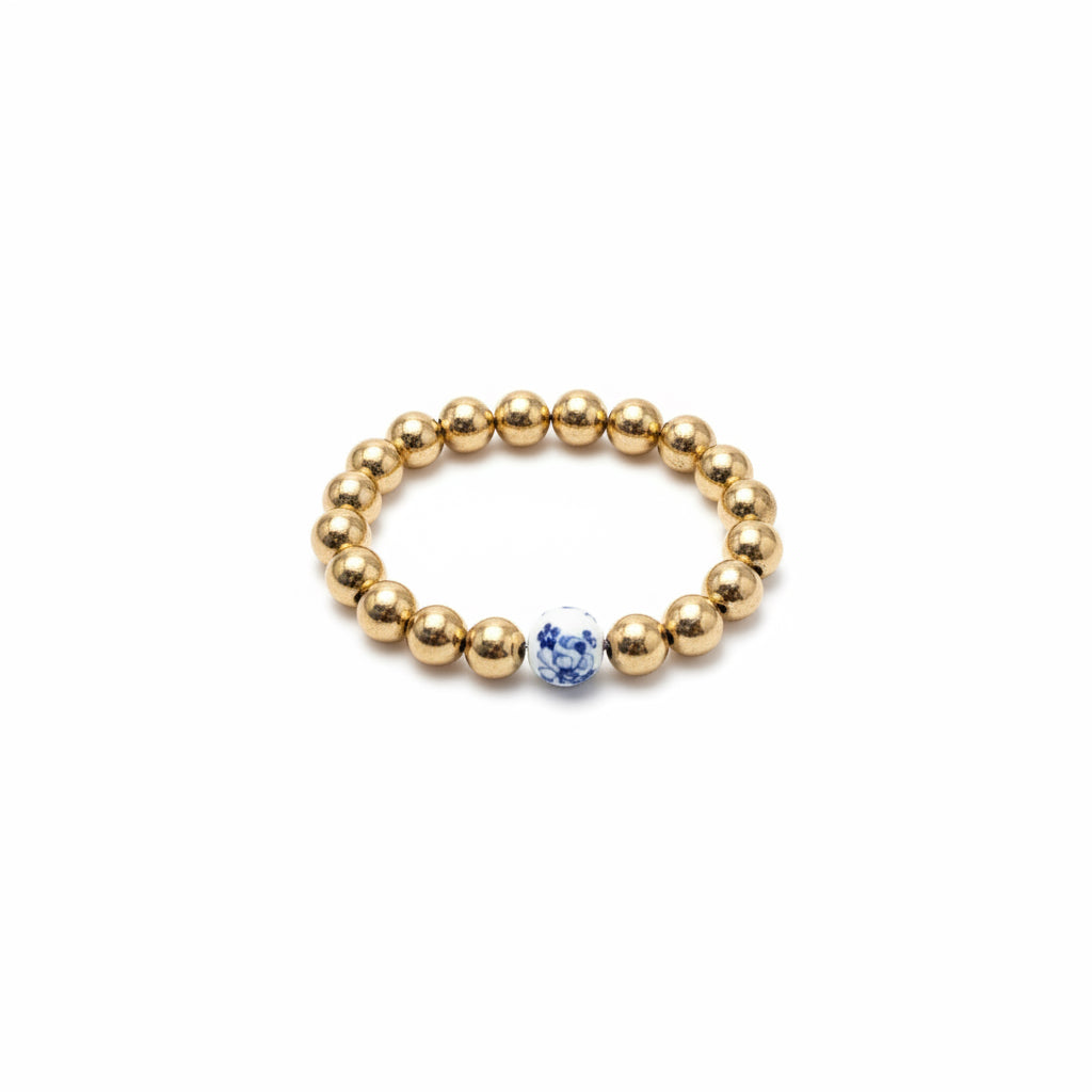 The Chinoiserie Pearl Stretch Bracelet has shiny gold beads with a single white bead featuring blue floral patterns. Available sizes: Small, Medium, Large.