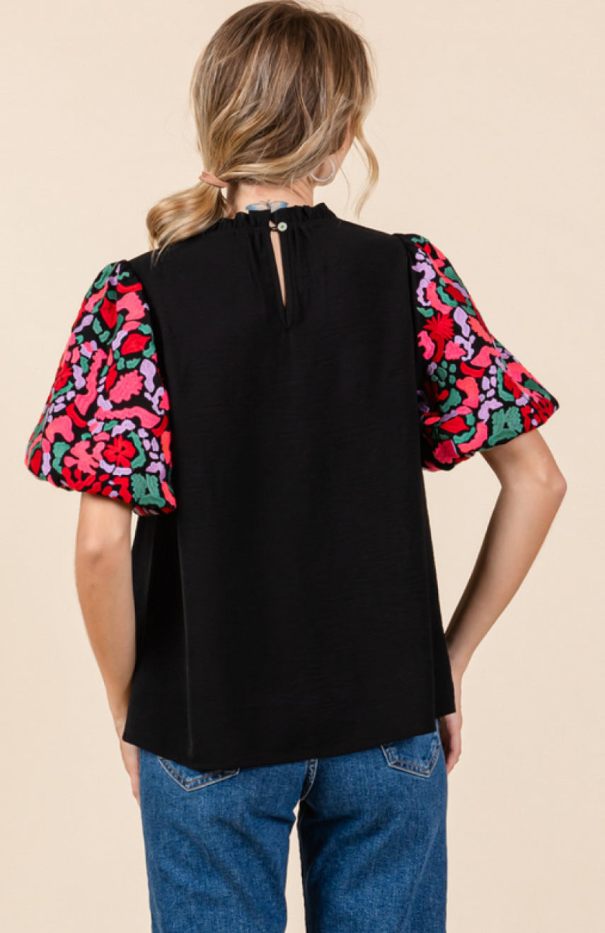 This is a black Frilled Floral Top with vibrant red and green floral print sleeves. Features include a single button closure at the neckline. Available in multiple sizes.