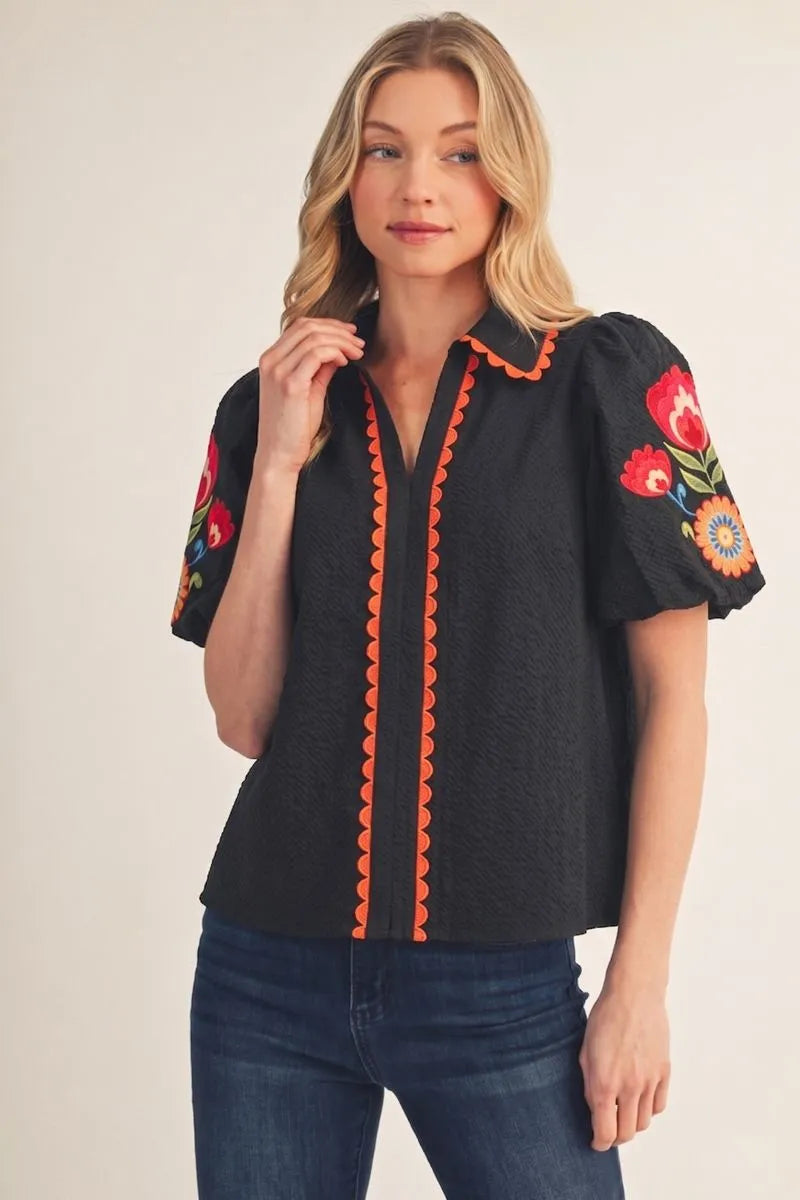 Fall in Bloom Blouse: black top with colorful floral embroidery on sleeves and orange scalloped trim. Available in sizes XS-XL.