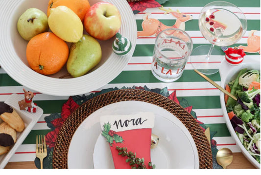Festive holiday table setting with a Baby Bowl Pinstripes (available in small and medium sizes) filled with assorted fruits, a plate with a "Nora" name card, petite red stocking, glass of bubbly drink, water glass, salad, cookies, and cutlery on a green and white striped tablecloth.