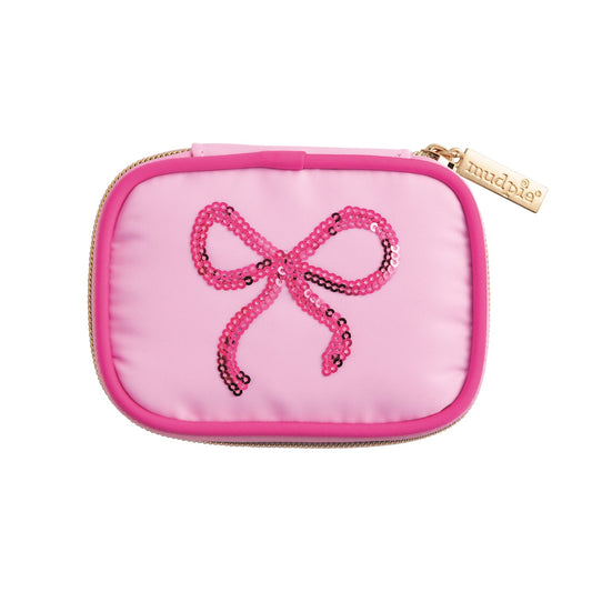 Small pink rectangular travel pill case with zipper, gold "mudpie" tag, sequined pink bow. Available in one size.