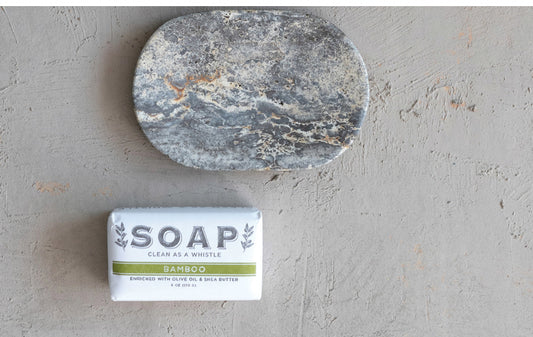 This product is a bar of bamboo soap labeled "clean as a whistle". It is accompanied by a round, grey stone with a textured surface. Both items are presented on an Oval Travertine Soap Dish located on a light grey concrete surface. The sizes available for this product have not been specified.