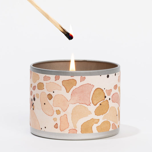 The Cashmere Kiss 4 oz. Tin Candle in a round tin with a pink and tan abstract design; available as a stylish travel candle.
