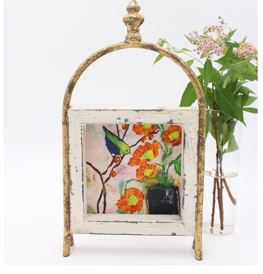This vintage-style 6x10 framed wood art on a metal stand captures a vividly painted canvas of a blue bird amidst orange flowers. It also has an accent feature - a glass vase filled with pink flowers set in the background.