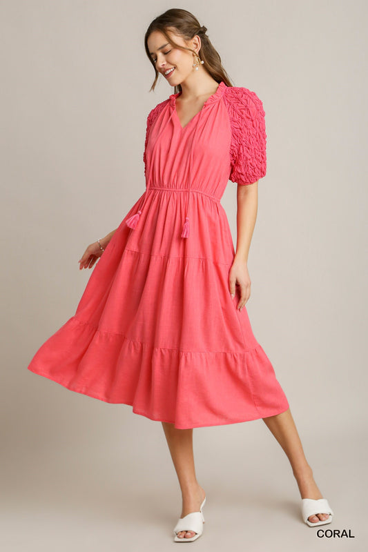 The Coral Linen Midi Dress features a v-neck, ruched sleeves, a cinched waist with tassel ties, and a tiered skirt. Available in sizes XS to XL.