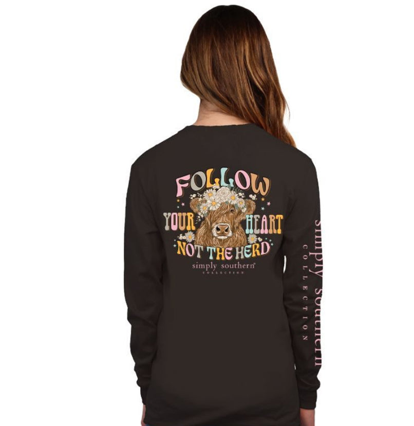 A woman with long brown hair, wearing a long-sleeve black shirt featuring the Herd Comet T-shirt logo and the phrase "FOLLOW YOUR HEART NOT THE HERD" alongside a graphic of a cow