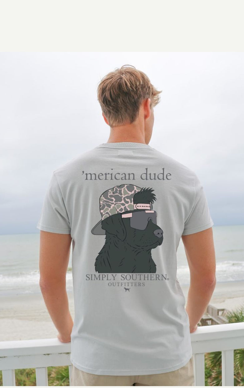 A man with blond hair wearing a Dude Whitewater TShirt stands facing a beach beyond a balcony railing.