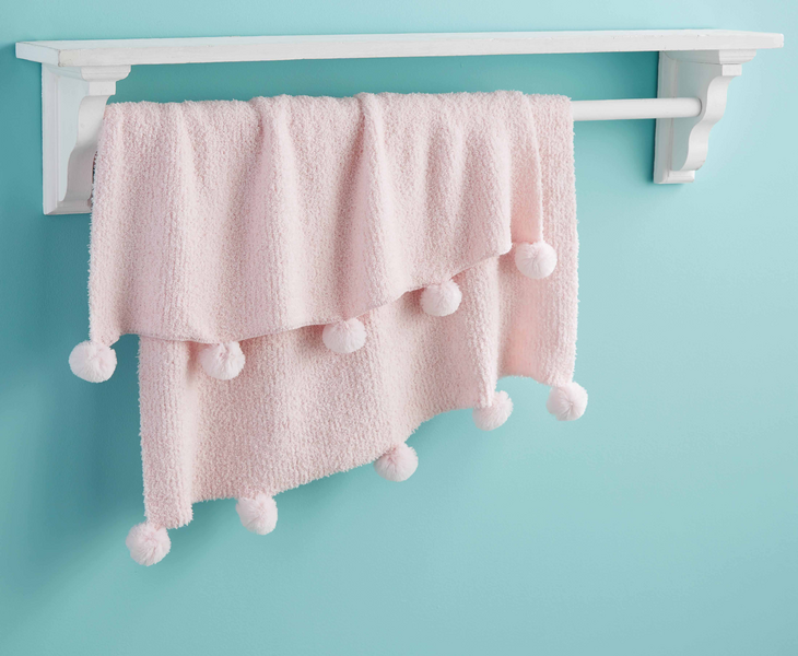 A soft Pink Chenille Blanket with white pom-poms hangs neatly on a white wooden rack, against a light blue wall.