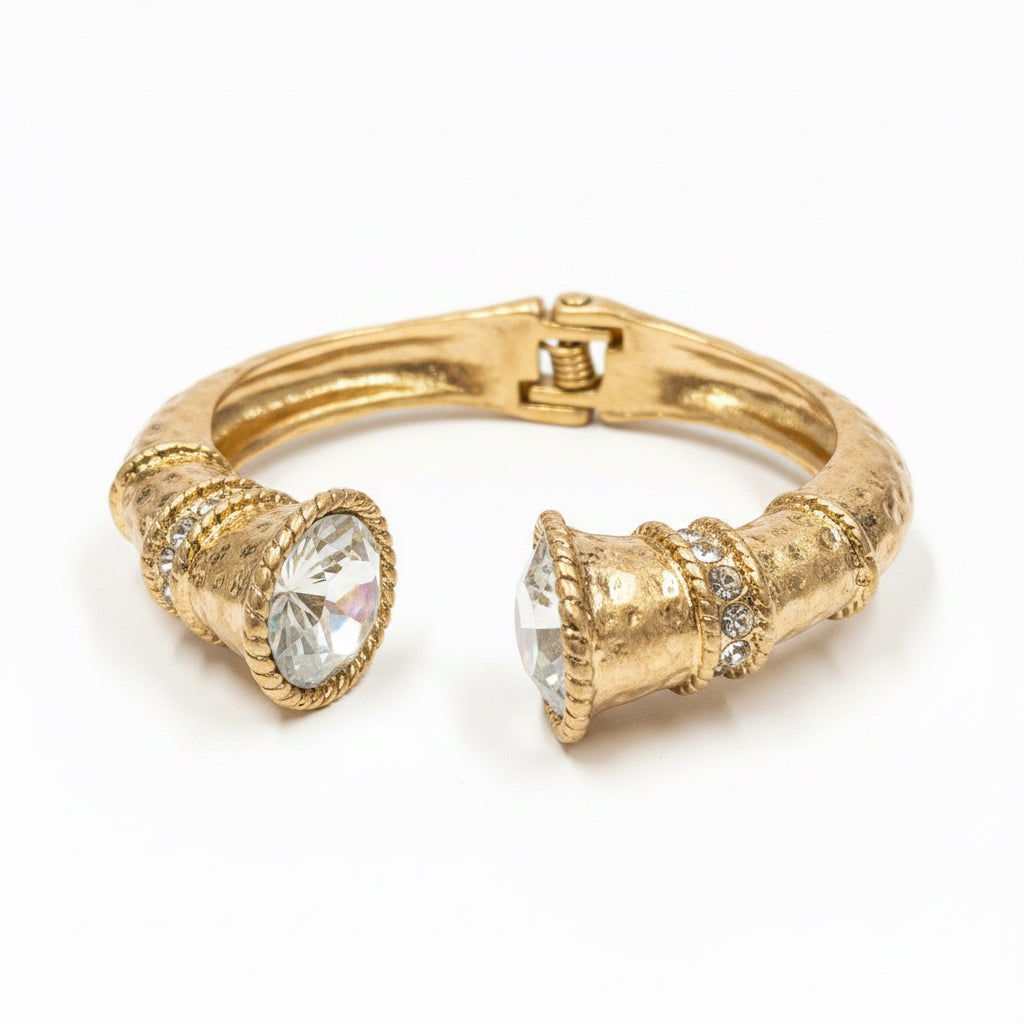 The Jojo Gold Clear Crystal Hinge Bangle features a gold textured design with two large, faceted clear crystals at each end, connected by a gold hinge. Available in multiple sizes.