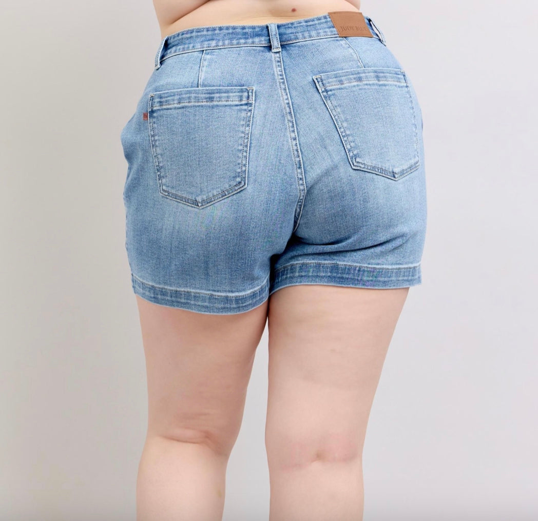 Judy Blue Mid Rise Trouser Shorts in light blue feature visible stitching, back pockets, and a leather patch on the waistband. Available sizes: XS to XL.