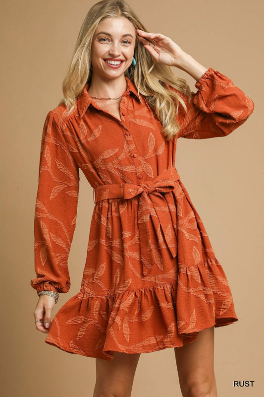 Piper Jacquard Dress: rust color, long sleeves, leaf pattern, ruffled hem, tie waist. Available in sizes XS–XL.