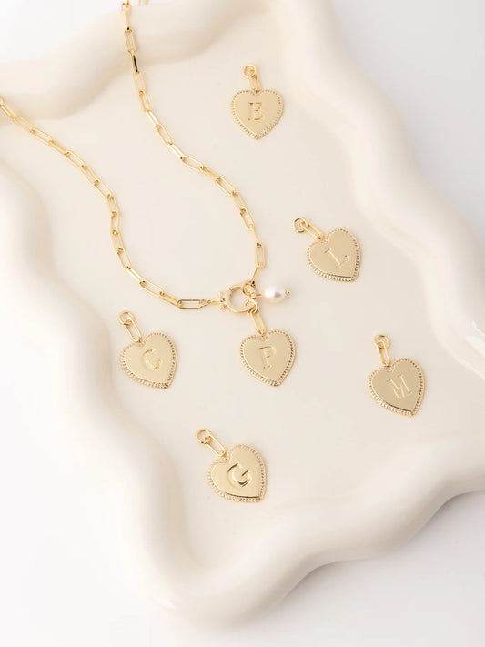 Thea Charm Bar’s customizable necklace features gold heart-shaped initial charms (E, L, M, G, C, F) on an 18k gold plated chain with a pearl accent. Hypoallergenic. Available in 16″, 18″, and 20″ lengths.