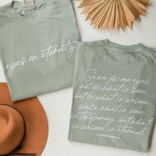 Two sage green Comfort Colors Christian tees with white script in Eyes on Eternity and 2 Corinthians 4:18. Sizes S-XXL.