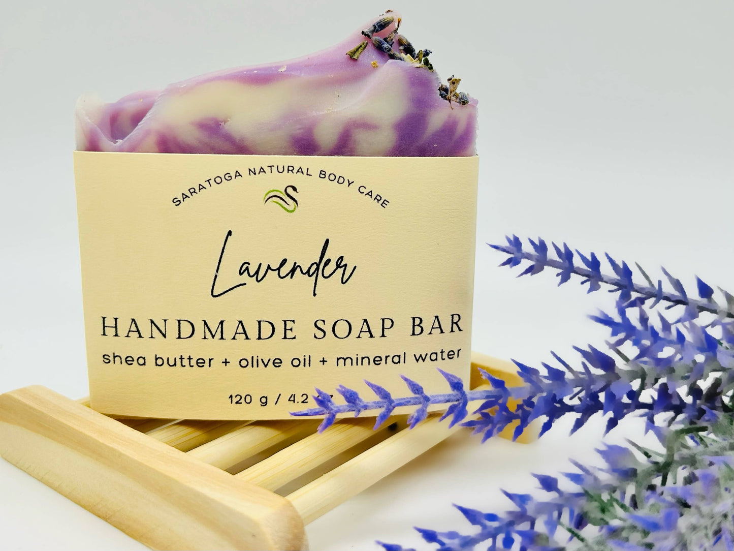 A Lavender Handmade Soap Bar from Saratoga Natural Body Care, VEGAN COLD PROCESS, boasts ingredients such as shea butter, olive oil, and mineral water. It comes with a wooden holder for easy storage and use. Sizes available may vary due to the handcrafted nature of the product.