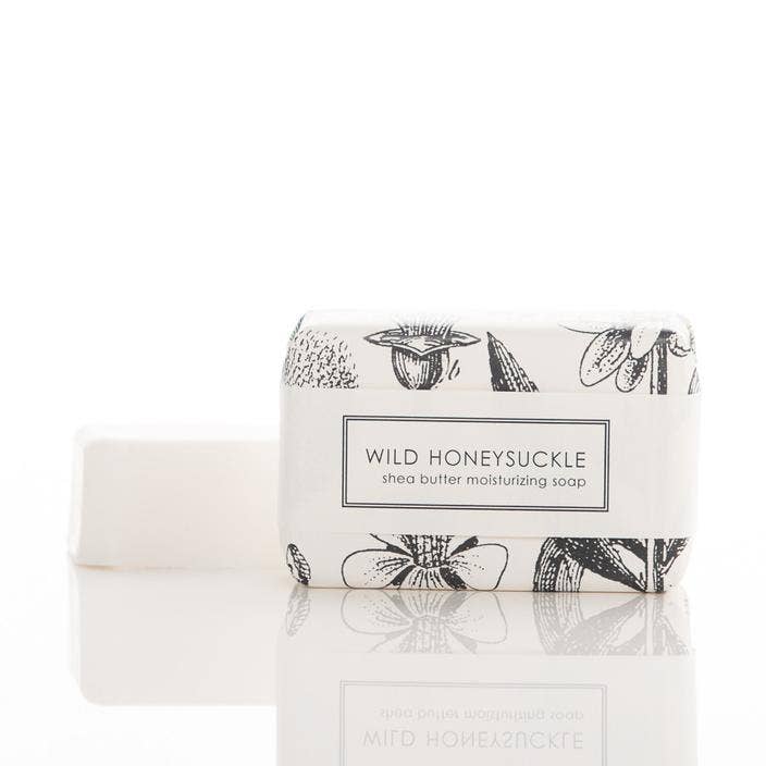 This product is a set of two bars of Wild Honeysuckle Soap - Bath Bar. The soap is made from shea butter, entirely vegan and features the aroma of wild honeysuckle. One bar comes unwrapped while the other is neatly packed in a package with floral designs. The soaps are placed on a reflective white surface for display purposes only. Available sizes not stated.
