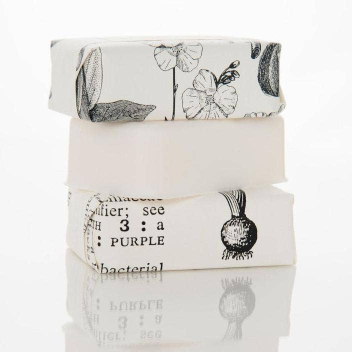 Gardenia Blossoms Soap - Bath Bar is a set of three stacked soap bars. Each bar is individually wrapped in paper featuring floral and botanical prints, with a prominent gardenia blossom design. The soap bars come attractively reflected on a white surface.