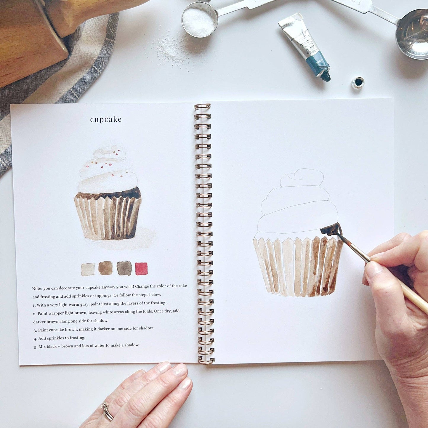 Baking Watercolor Workbook, spiral-bound beginner book (sizes: small, medium), shows painted cupcakes and color swatches.
