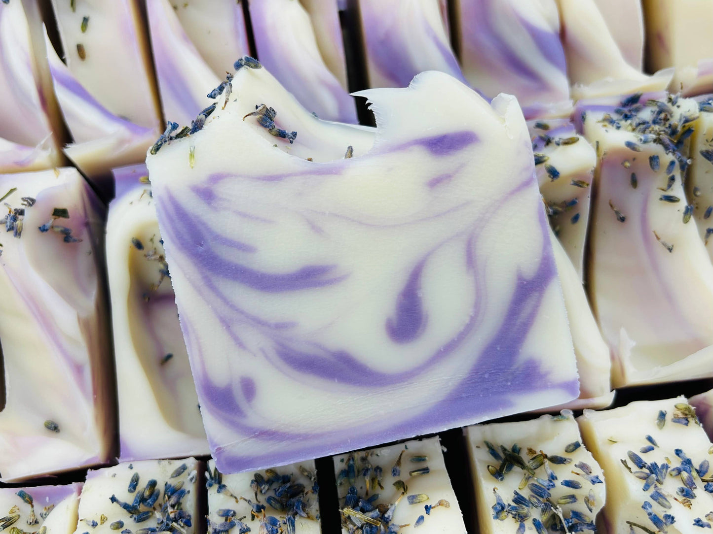 This is a collection of Lavender handmade soap bars, characterized by unique swirls of purple and white colors. Each bar is embellished with a sprinkling of dried lavender on top for additional fragrance and aesthetic appeal. These soaps are displayed in neat rows and available in standard bath size.