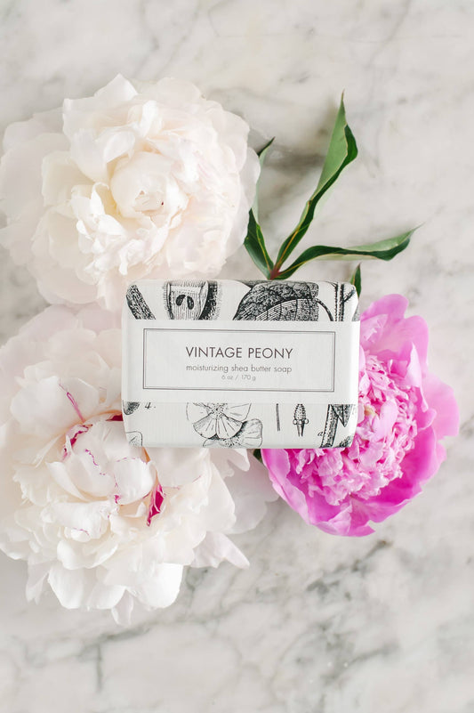This Vintage Peony Soap is a bath bar presented in beautiful floral paper wrapping. It carries the delicate scent of lush white and pink peony flowers. The soap has been crafted to capture the aesthetic of a vintage era, resting against an elegantly polished marble surface. The product is available in standard soap bar size.