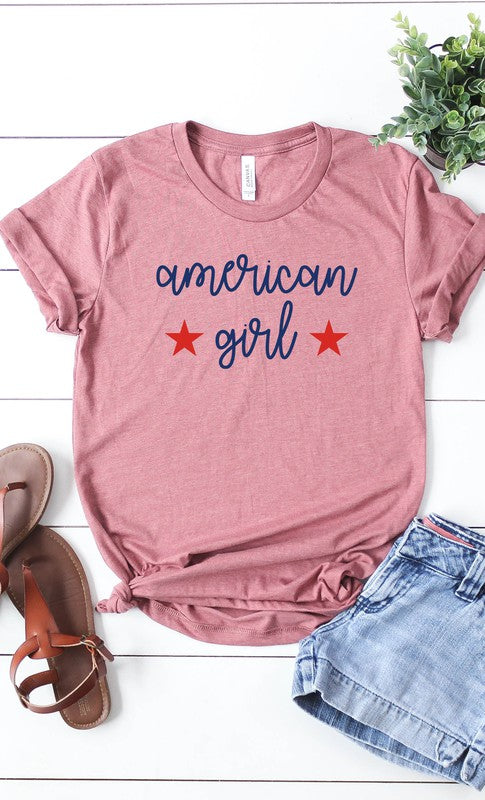 A Heather Mauve American Girl Tshirt with the phrase "american girl" printed in blue lowercase letters, flanked by two red stars, laid flat next to denim shorts and brown sandals on a wooden surface