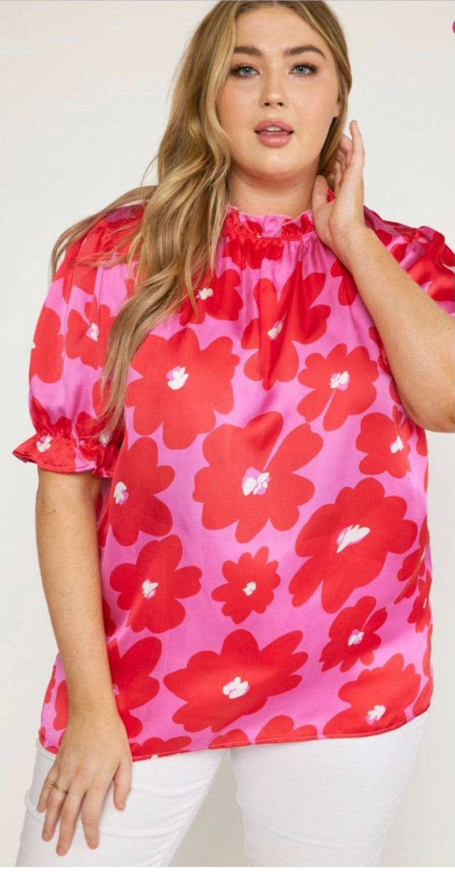 A woman in a Poppy Ruffle Blouse with white pants is sizing her neck gently while looking towards the camera.
