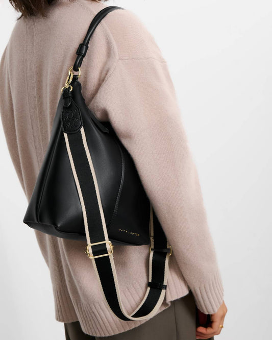 Hallie Satchel Bag in black vegan leather with gold hardware, embossed logo near the top, and a wide black and white shoulder strap; available in small, medium, and large sizes.