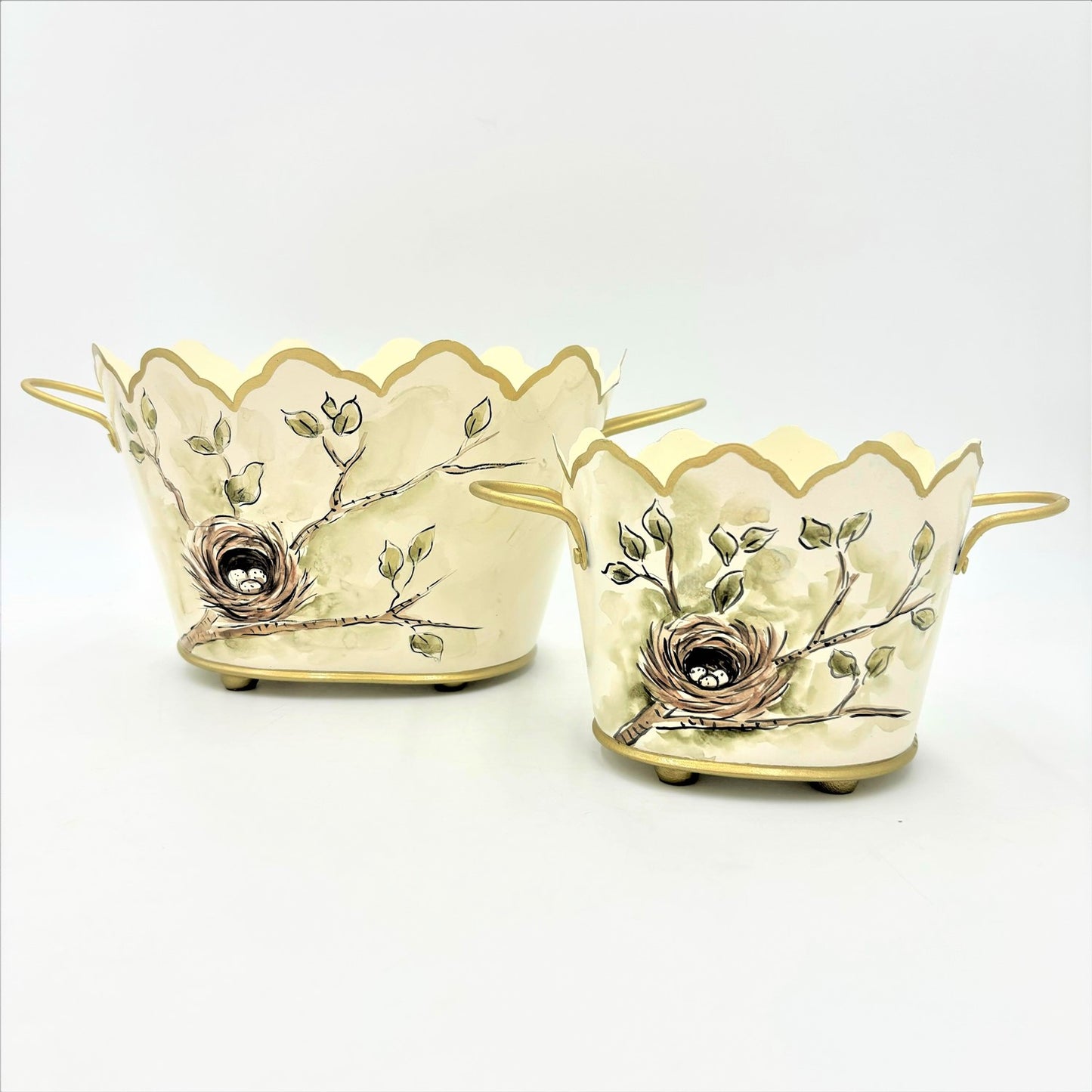 The Handpainted Scalloped Metal Cachepots, Bird's Nest are cream-colored metal containers with handles, scalloped edges, and bird nest designs featuring silver-toned eggs. Available in two sizes: 8" and 10".
