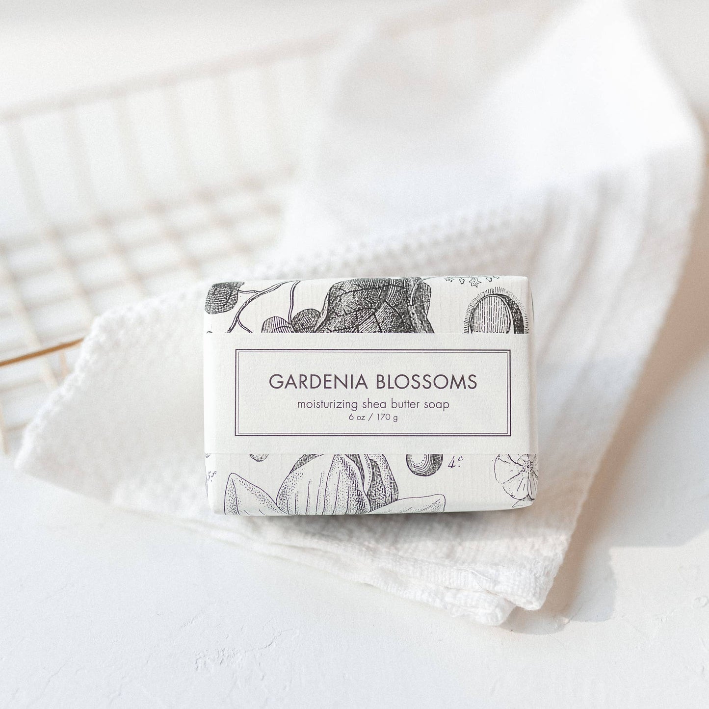 The Gardenia Blossoms Soap is a Bath Bar enveloped in a paper wrapper featuring a floral design. The soap offers a refreshing scent derived from gardenia blossoms. It boasts an aesthetic appeal with its rested placement on a textured white towel, providing it with clean and simple looks. The size of the product varies depending on availability.