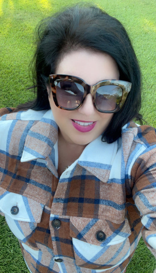 A selfie of a woman with dark hair wearing large sunglasses and a Brushed Plaid Button Down Shirt - PLUS, smiling while lying on green grass.