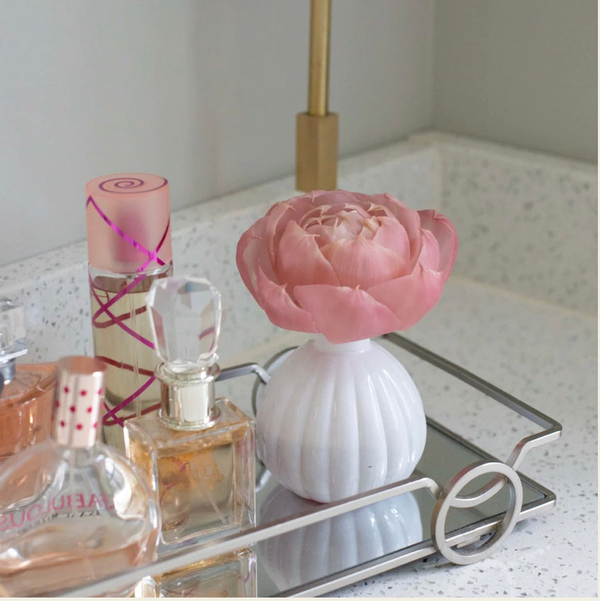 The product is a Sweet Grace Flower Diffuser BW171125. It features an aromatic bouquet resting on a mirrored tray alongside a white ceramic vase containing a pale pink, artificial peony flower. The set-up is designed to be placed on a marble bathroom counter. The perfume bottles within the collection vary in size.