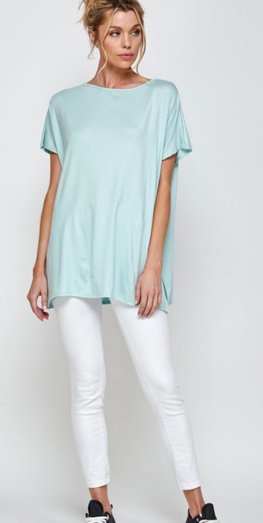 A woman stands wearing a light aqua blue, buttery soft A.gain “The Perfect” Short Sleeve Shirt Plus paired with tight white pants and black slip-on shoes, posing against a plain background.