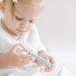 A toddler with light hair and a white headband examines a Pacifier Clip - Pink and Blue attentively in a brightly lit, neutral-colored room.
