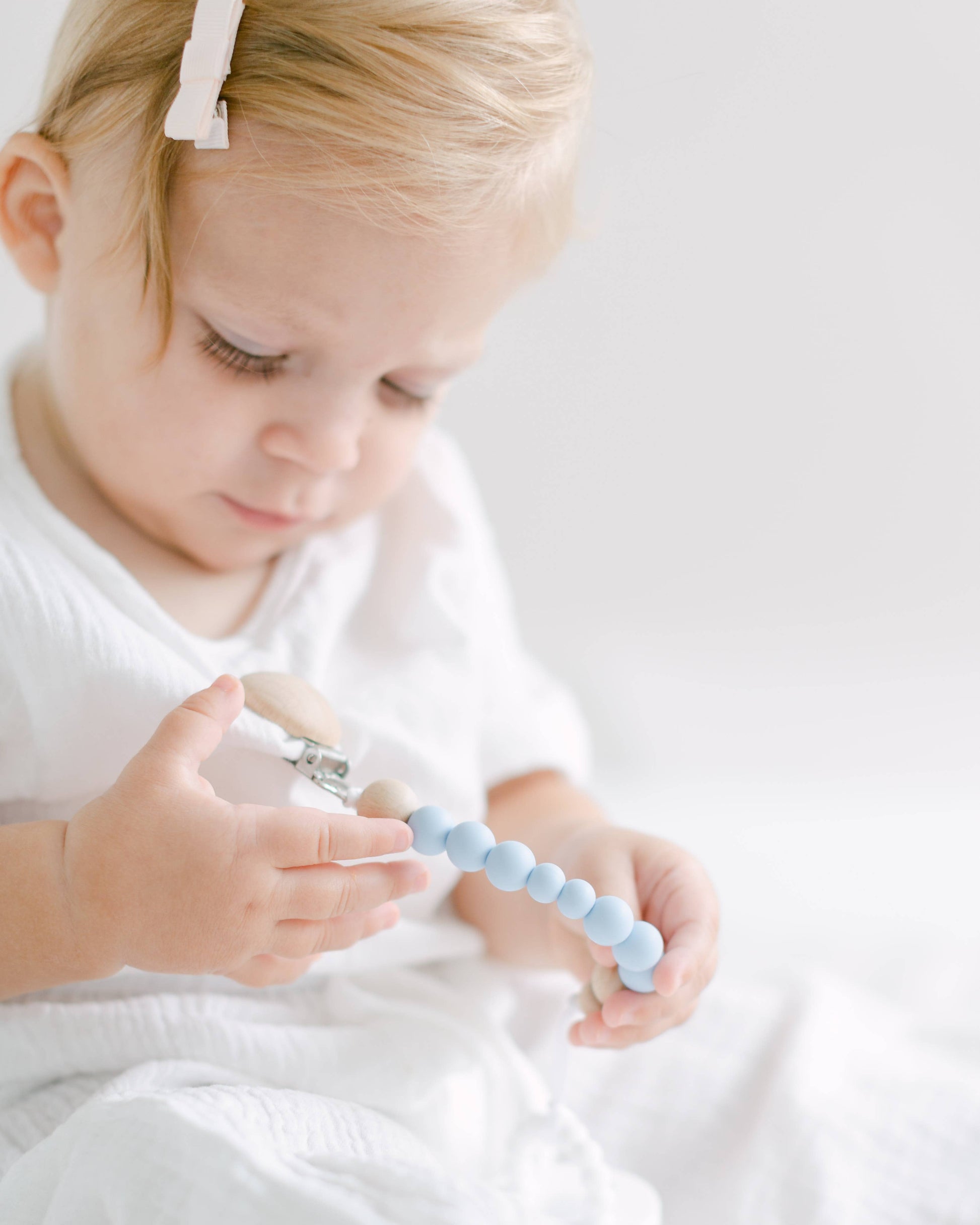 A toddler with light hair and a white headband examines a Pacifier Clip - Pink and Blue attentively in a brightly lit, neutral-colored room.